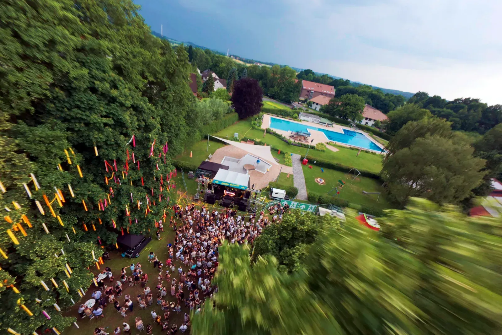 FPV-Flug über das Hille Beatz Open Air 2025: geschmückte Bäume, Bühne und feiernde Menge, dahinter das Freibad Dörentrup