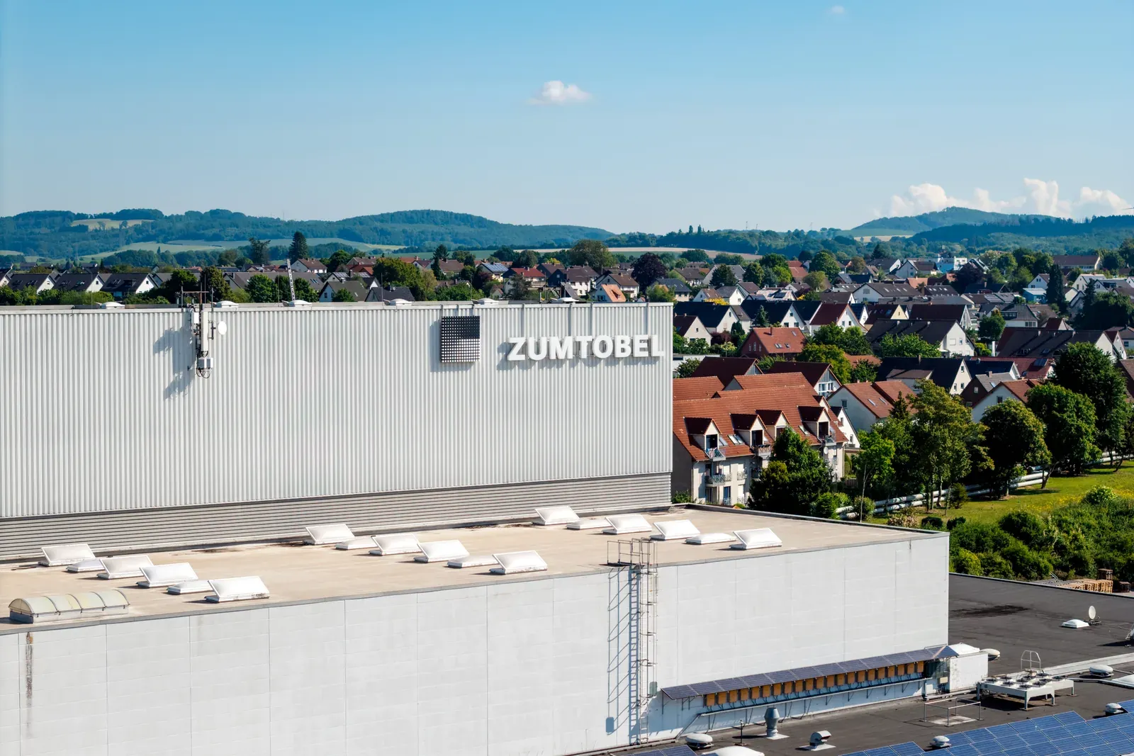 Zumtobel-Werk in Lemgo aus der Luft: Hallenfassade mit Logo, dahinter Wohngebiet und Hügelland